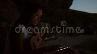 嬉皮青年女子晚上在海滩上使用她的手机学习如何演奏四弦琴吉他。 1920x1080